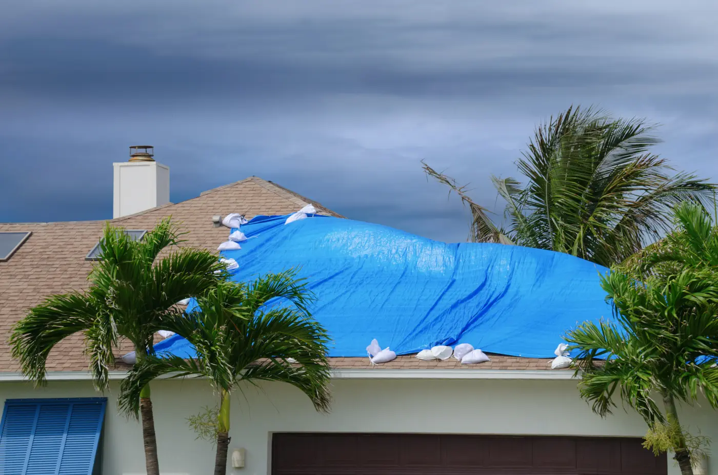 Storm-damaged residential roof covered with a blue tarp and sandbags, surrounded by palm trees.